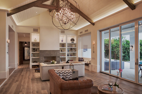 A living room lit in layers — cove light washing the vaulted ceiling, an orb chandelier, sconces, lit shelving and a shade concealed above the window.